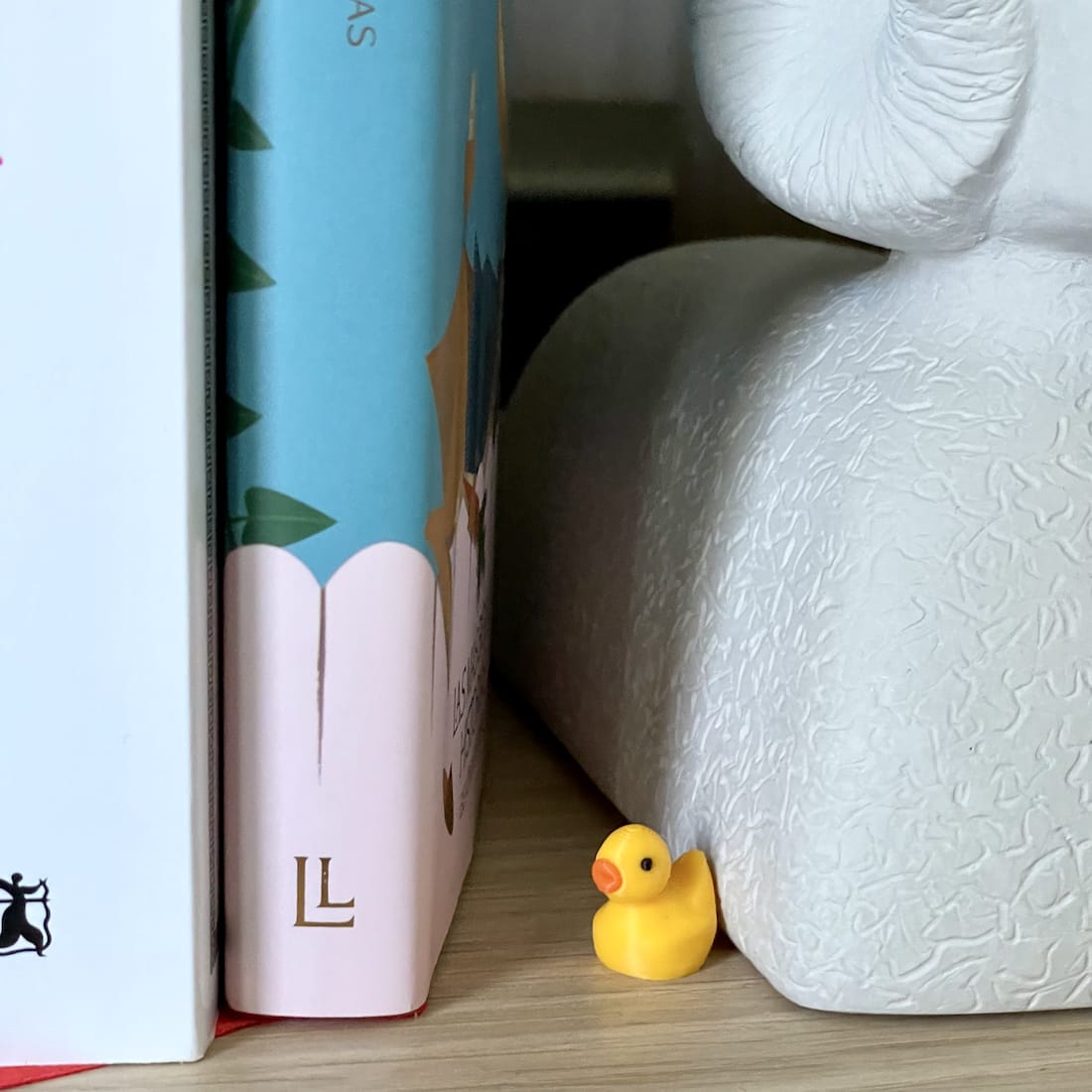 Single tiny yellow duck hidden between books and a decorative figure on a shelf