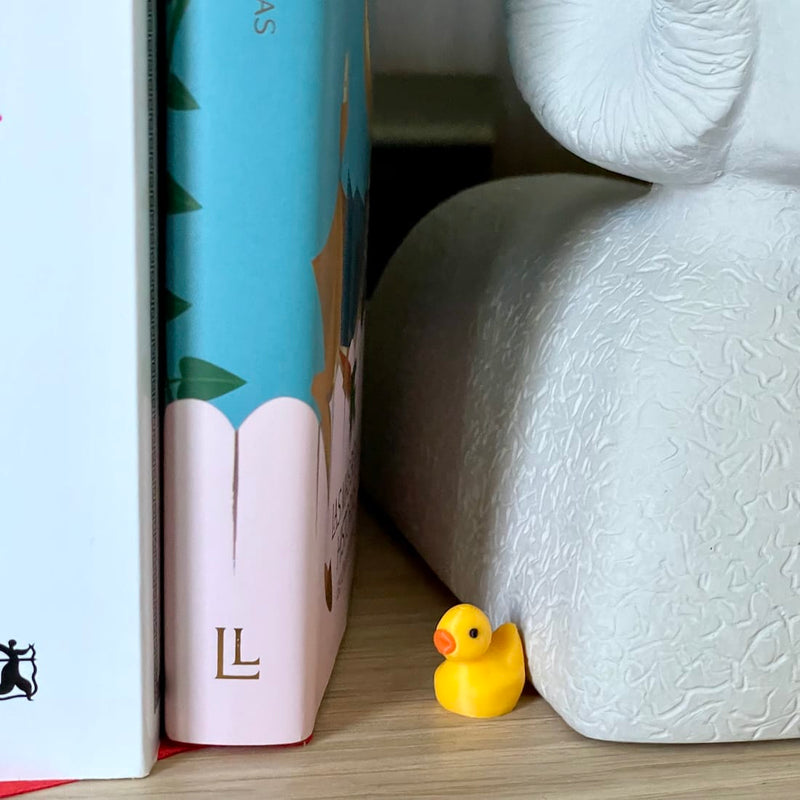 Single tiny yellow duck hidden between books and a decorative figure on a shelf