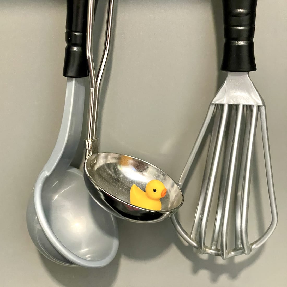 Tiny yellow duck hidden inside a kitchen ladle hanging next to a whisk