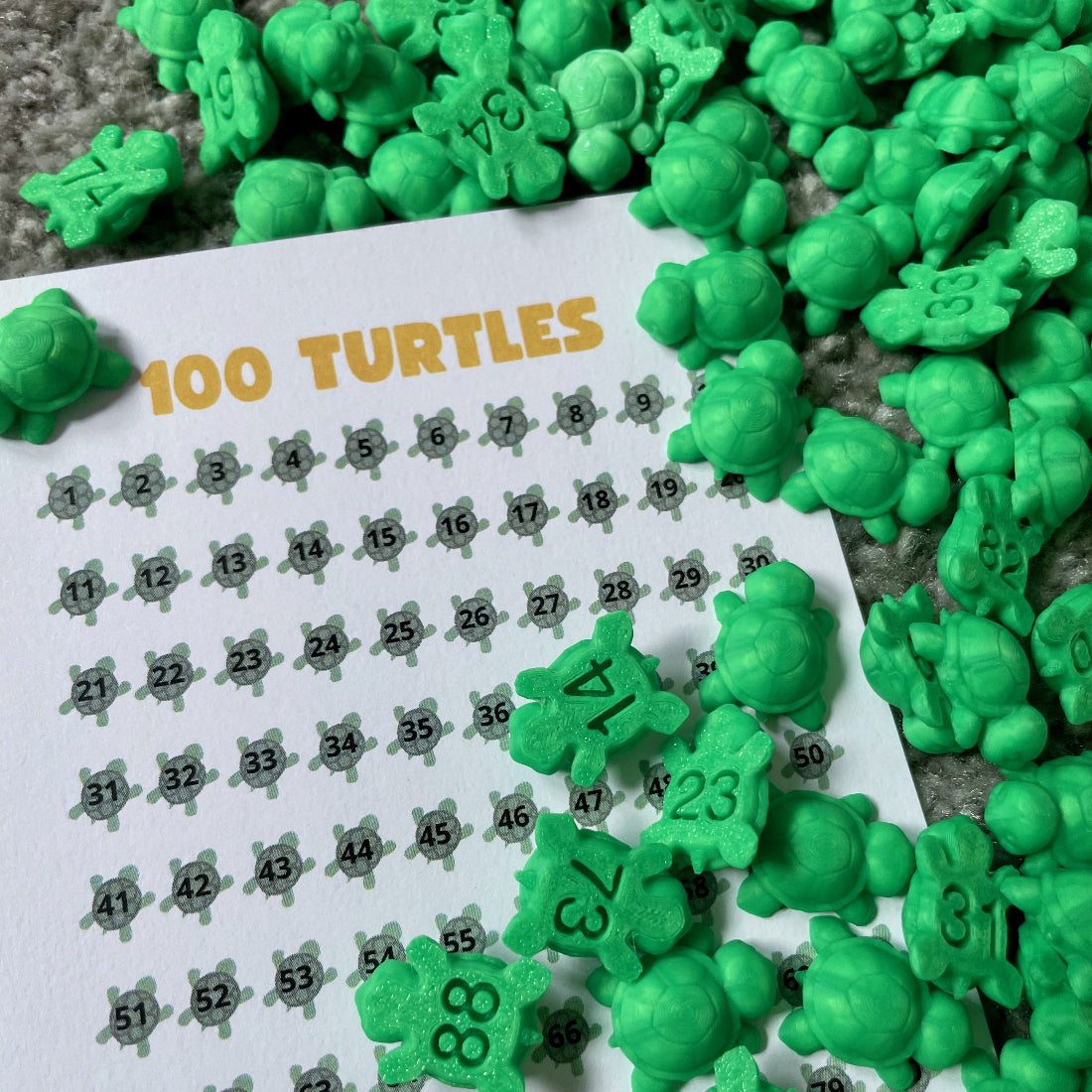 100 small green numbered turtles spread out next to a 100 Turtles tracking sheet